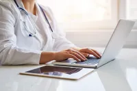 Doctor wearing white coat typing on laptop with tablet on white desk under bright window light.