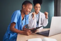 Two female medical professionals smiling and celebrating while looking at a tablet and laptop in an office.