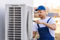 Technician in blue overalls and cap repairing an outdoor air conditioning unit with a screwdriver.