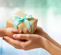 Adult hands gently holding a small gift wrapped in brown paper with a light blue ribbon from a child's hands.