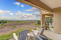 Two Adirondack chairs on a beachside patio overlooking dunes and a partly cloudy blue sky.