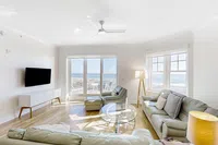 Bright modern living room with light gray sofas, glass coffee table, and ocean view through large windows.