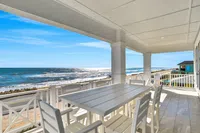 Spacious beachside patio with white dining table and chairs overlooking sunny ocean waves and sandy shore.