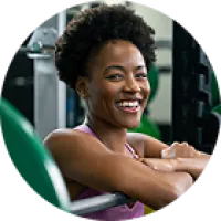Smiling woman with curly hair in purple top resting on gym equipment in fitness center.