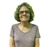 Smiling elderly woman wearing glasses, a purple V-neck T-shirt, and dark pants, standing with hands clasped.