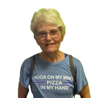 Smiling elderly woman with glasses wearing a blue t-shirt that says Yoga on my mind, pizza in my hand.