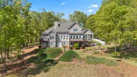 Large gray multi-story house with deck surrounded by lush green trees under blue sky on sloped land