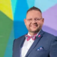 Smiling man in a blue suit and pink bow tie stands before a colorful geometric wall mural.