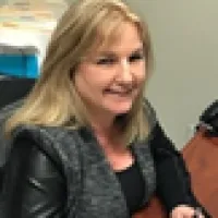 Smiling woman with blonde hair wearing a gray vest and black top sitting at a desk indoors.