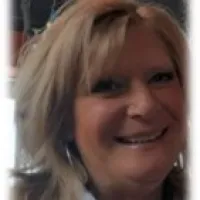 Smiling middle-aged woman with blonde hair wearing a white jacket and dark top indoors.