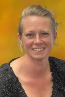 Smiling woman with blonde hair wearing a black polka dot blouse against a warm yellow background