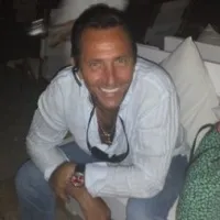 Man smiling wearing white shirt and blue jeans sitting on a chair with sunglasses hanging from collar.