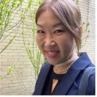 Smiling woman with pearl necklaces and navy blazer standing outdoors by light brick wall and green plants.