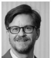Black and white portrait of a smiling man with glasses and beard in a suit jacket