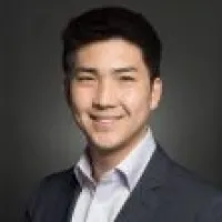 Smiling young man with short dark hair in a dark suit jacket and white shirt against a gray background.