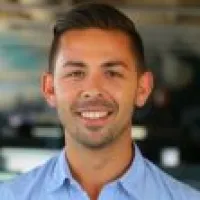Smiling young man with short dark hair wearing a light blue shirt in an office environment.