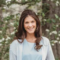 Smiling woman with long brown hair wearing light blue top and white knitted cardigan outdoors near blooming trees