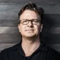 Middle-aged man with glasses and dark shirt poses against a dark wooden plank background.