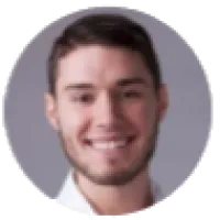 Smiling young man with beard and short brown hair against a neutral gray background.