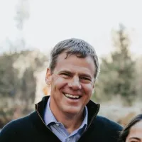 Smiling middle-aged man in casual blue shirt and black jacket outdoors with blurred trees in background