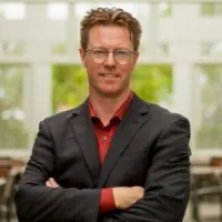 Smiling man in a blazer stands confidently with arms crossed in a bright indoor space.
