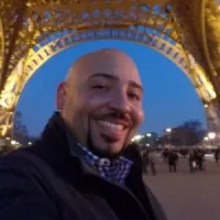 A smiling man takes a selfie under the illuminated Eiffel Tower at dusk, capturing a cheerful moment.