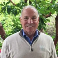 Smiling man in a light sweater stands against a lush green background with flowering plants.