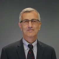 A professional man with glasses, dressed in a suit, smiling against a neutral backdrop.
