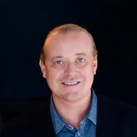 Smiling man in a black blazer against a dark backdrop with soft lighting.