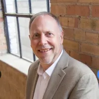Middle-aged man smiling in a beige blazer and white shirt by a window with brick wall background