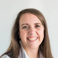 Smiling woman with long brown hair wearing a white shirt and black vest against a plain light background
