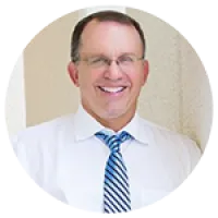 Smiling middle-aged man wearing glasses, white shirt, and blue striped tie against a neutral background
