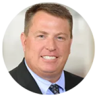 Smiling middle-aged man in suit and blue shirt with patterned tie against neutral background.
