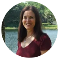 Smiling woman with long brown hair wearing a maroon top standing outdoors near a lake with trees in background