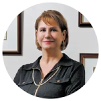 Middle-aged woman with short light brown hair wearing a dark blouse, smiling against a white wall with framed pictures.