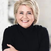 Smiling middle-aged woman with short blonde hair wearing a black turtleneck sweater and hoop earrings.