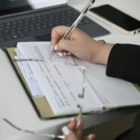 Person writing notes in a notebook beside a laptop and smartphone on a white desk.