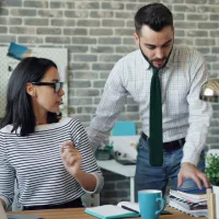 Two colleagues discussing work at a desk with laptop, notebooks, and plants in a modern office space.