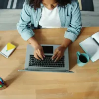 Overhead view of a person typing on a laptop at a wooden desk with headphones, notebook, coffee, and plant.
