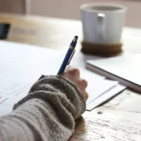 Person writing with blue pen on paper at wooden desk with notebook and coffee mug nearby