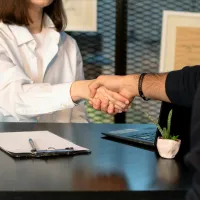 Two professionals shaking hands over a desk with laptop, clipboard, and small plant, symbolizing agreement.