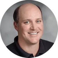 Smiling middle-aged man with light skin, blue eyes, and short brown hair wearing a black polo shirt on gray background
