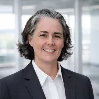Professional middle-aged woman in a dark blazer and white shirt smiling with an office background.