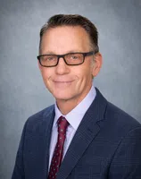 Professional middle-aged man wearing glasses, a blue checkered suit, light purple shirt, and patterned red tie smiling against gray background