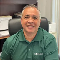 Smiling man in green SRI Healthcare polo shirt sitting in office with papers and printer in the background