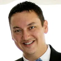Close-up of a smiling man with short dark hair wearing a black suit and blue patterned tie against a white background