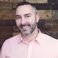 Smiling man with short dark hair and beard wearing light pink shirt against wooden wall background.