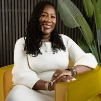 Smiling woman with long curly hair wearing white dress seated on yellow chair with green plant background