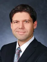 Professional man in a business suit with a patterned tie smiling against a dark gray background