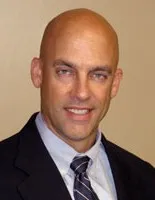 Professional headshot of a bald man in a dark suit with a striped tie smiling against a plain background